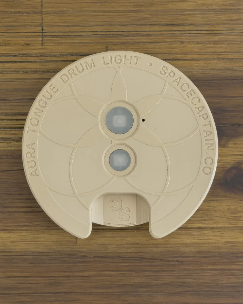 The control face of the Aura Light on a wood table, engraved "Aura Tongue Drum Light · spacecaptain.co" around two buttons, with a USB-C port at the edge